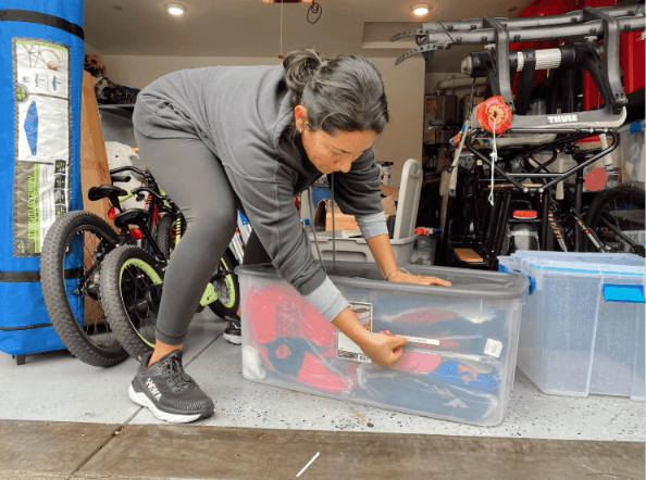 Woman in a grey hoodie labels a clear storage bin in a cluttered home garage.